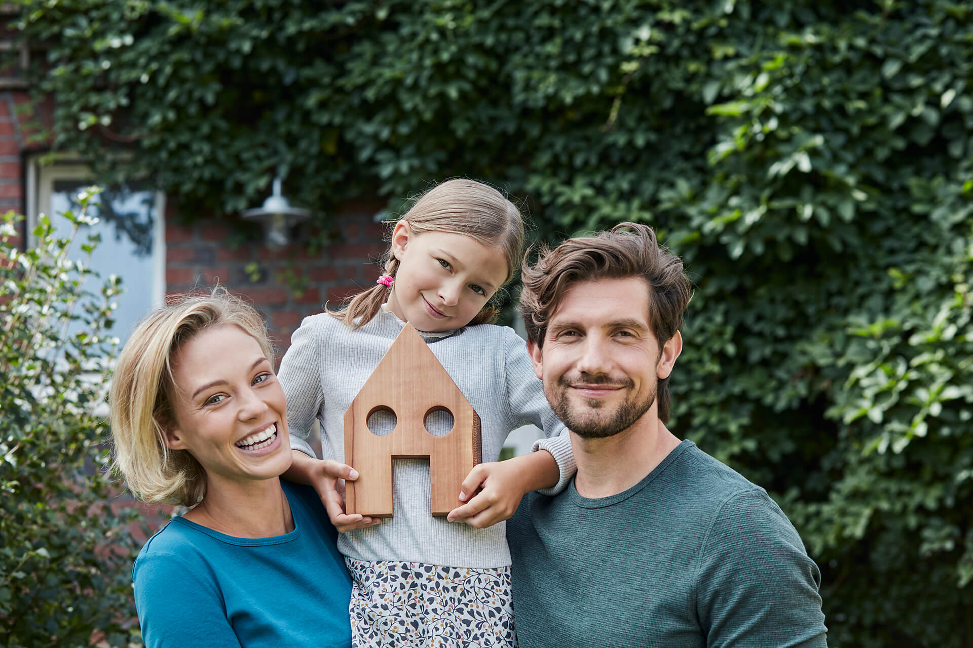 Familj framför sitt nya hus; dottern håller ett litet trähus i händerna som symbol för deras nya hem.